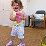toddler, child, smiling, indoor, sippy_cup, curly_hair, colorful_clothing, shoes, wooden_floor, chair, cushion, cabinet, heater, happy, person, shorts, tank_top, playful, expression, standing
