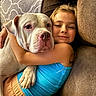 child, dog, hugging, couch, blanket, indoor, smiling, white_dog, blue_tank_top, brown_couch, comfort, pet, friendship, relaxation, home, happy, closeup, face, arm, casual_clothing