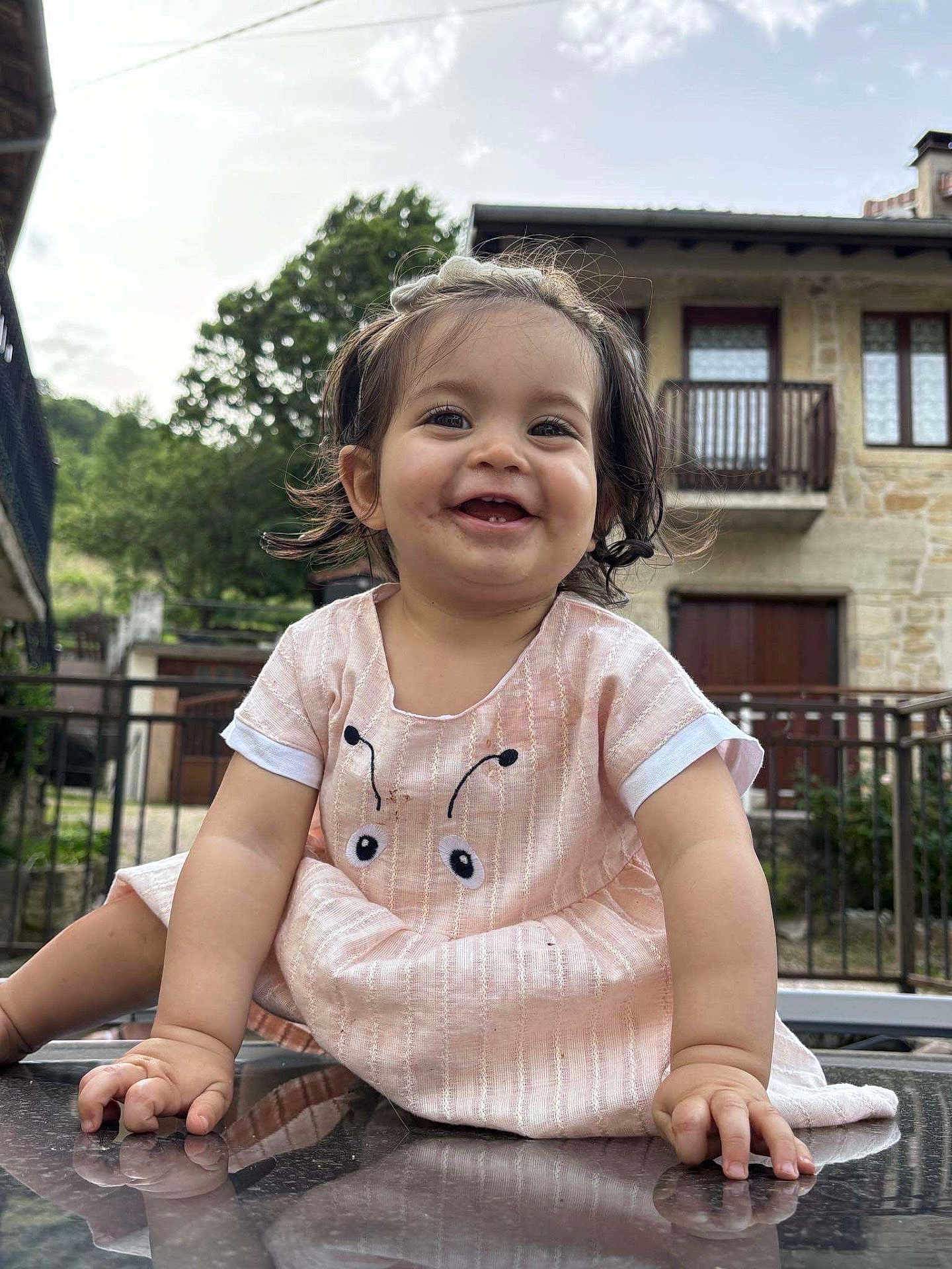 Lily-Ana participe au concours pour gagner de l'argent avec cette photo : toddler, child, smiling, outdoor, dress, reflection, hands, hair, pigtails, face, happy, building, window, railing, nature, tree, sky, stone_wall, person, candid