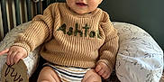 Ashton is registered to the contest to win money with this photo: baby, child, infant, sweater, knitted_clothing, striped_diaper, quilt, cushion, wooden_sign, five_months, bed, crib, camera, indoor, person, cute, smiling, face, skin, hand
