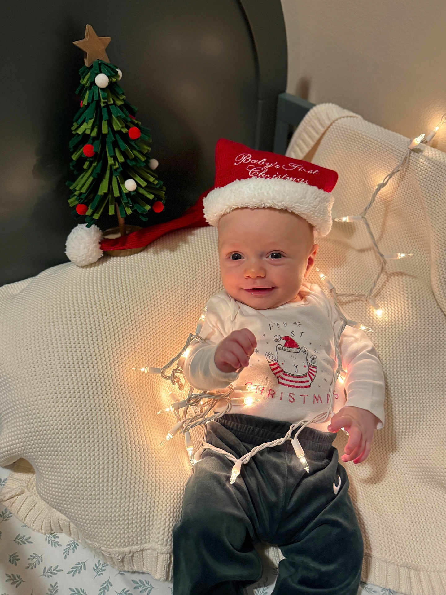Ashton is registered to the contest to win money with this photo: baby, child, christmas, holiday, festive, santa_hat, string_lights, smile, cute, cozy, blanket, decor, bed, pajamas, indoors, happy, face, toy_tree, infant, warm