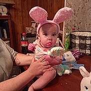 Oaklynn is registered to the contest to win money with this photo: adult_hand, baby, bottle, bunny_ears, child, clothing, cute, facial_expression, furniture, holding, indoor, lollipop, person, pink, plush_toy, table, tongue, toy, unicorn_toy, wood_paneling