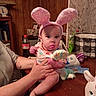 adult_hand, baby, bottle, bunny_ears, child, clothing, cute, facial_expression, furniture, holding, indoor, lollipop, person, pink, plush_toy, table, tongue, toy, unicorn_toy, wood_paneling