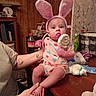 Oaklynn is registered to the contest to win money with this photo: adult_hand, baby, bunny_ears, child, colorful, curious_expression, cute, face, holding, household_items, indoor, lollipop, person, pink_clothing, scale, sitting, supporting_hand, table, toy, wood_paneling