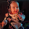 baby, child, smiling, headband, floral_pattern, clothing, hand_holding, indoors, portrait, cute, happy, infant, person, warm_lighting, seated, long_sleeves, necklace, fabric, blurred_background, skin