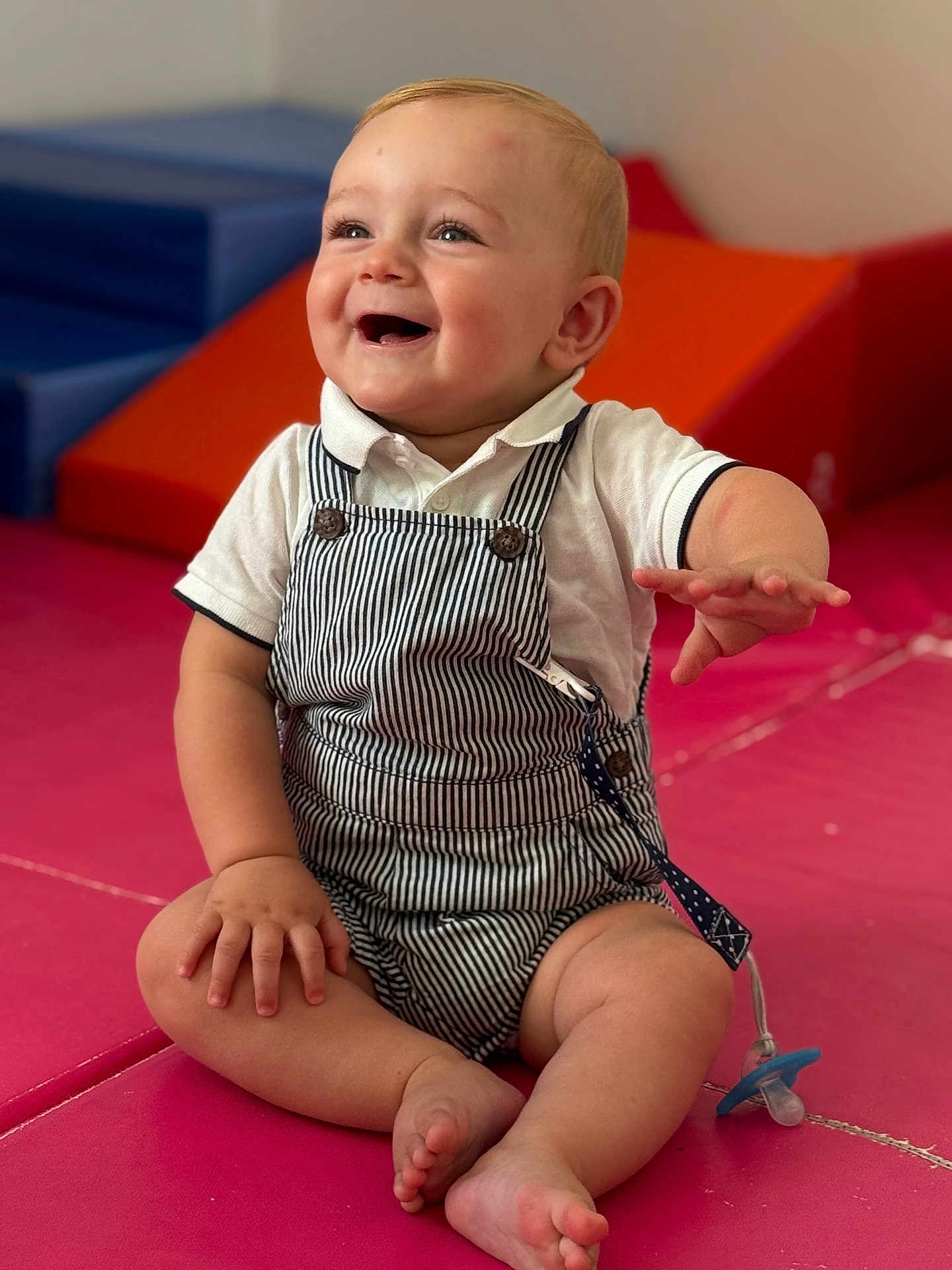 Giulian a rejoint le concours — aidez-le/la à gagner de superbes lots ! baby, child, smiling, sitting, overalls, white_shirt, play_mat, indoor, happy, cute, infant, person, face, hand, foot, toy, pacifier, striped_clothing, play_area, soft_surface