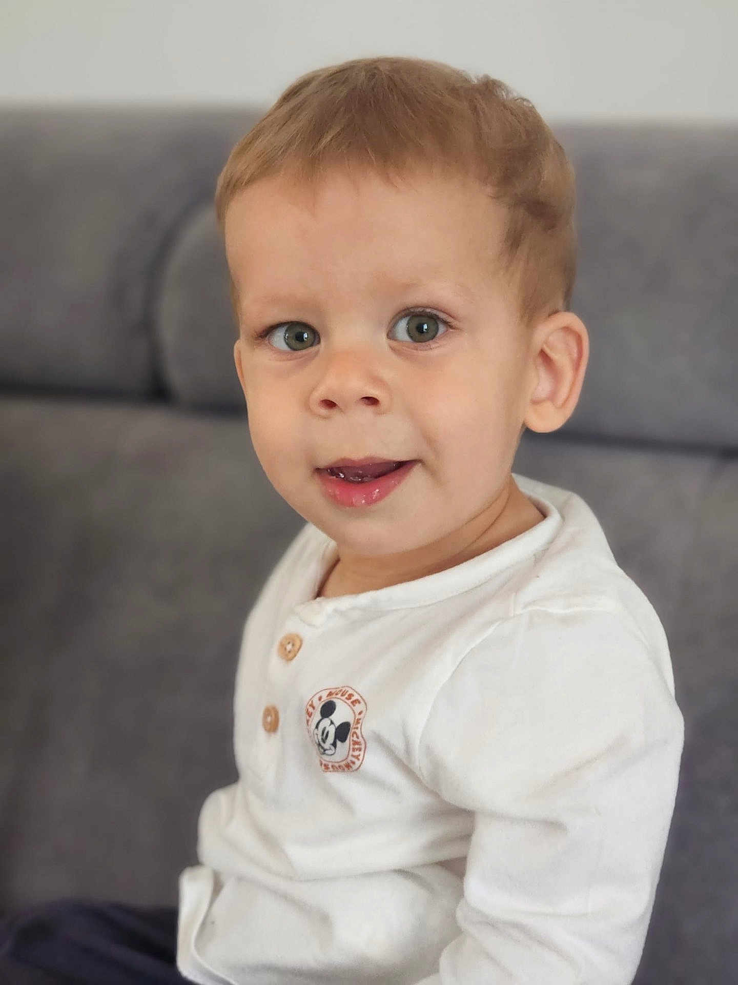 Noah participe au concours pour gagner de l'argent avec cette photo : toddler, child, face, smile, white_shirt, mickey_mouse, indoor, couch, seated, curious, young_child, portrait, casual_clothing, light_brown_hair, happy, cute, person, baby, head, eyes