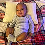 baby, infant, smile, pillow, blanket, plaid, clothing, onesie, socks, bed, comfort, lying_down, cute, indoor, child, person, happy, resting, young, cozy