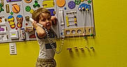 Ryder is registered to the contest to win money with this photo: toddler, child, toy_phone, barefoot, carpet, yellow_wall, activity_board, interactive_toys, playroom, curly_hair, shorts, tshirt, indoor, young_child, colorful, educational_toys, standing, expression, play, learning
