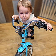 Ryder joined the competition — help win amazing prizes! toddler, child, bicycle, blue_bike, handlebars, indoor, wooden_floor, focused, barefoot, short_sleeves, blond_hair, young_child, toy, motion, play, flooring, metal, pedals, wheel, grip