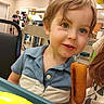 Ryder is registered to the contest to win money with this photo: toddler, child, corndog, food, high_chair, indoor, party, balloons, people, smile, face, blue_shirt, curly_hair, plate, table, adult, background, celebration, event, happy