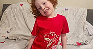 Kenji is registered to the contest to win money with this photo: child, red_hair, smile, red_tshirt, dinosaur_graphic, jeans, couch, blanket, heart_decorations, indoor, happy, posing, portrait, casual_clothing, home, furniture, curly_hair, person, kid, cozy