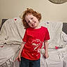 child, red_hair, smile, red_tshirt, dinosaur_graphic, jeans, couch, blanket, heart_decorations, indoor, happy, posing, portrait, casual_clothing, home, furniture, curly_hair, person, kid, cozy