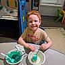 Kenji joined the competition — help win amazing prizes! child, boy, red_hair, painting, egg, paintbrush, green_paint, yellow_paint, table, classroom, art_supplies, smiling, sitting, plastic_bowl, spoon, chair, wall_art, window, carpet, indoor