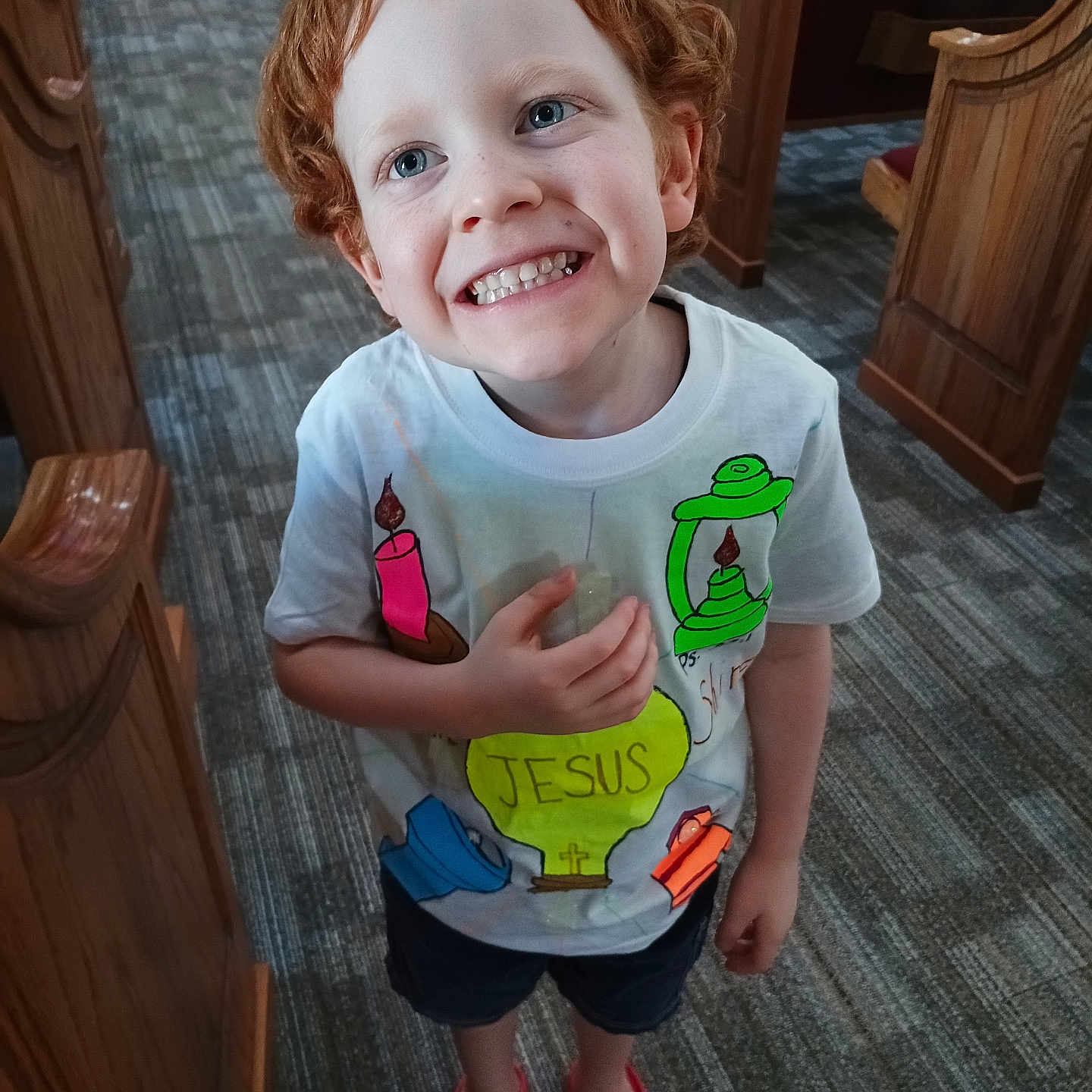 Kenji is registered to the contest to win money with this photo: child, red_hair, smiling, tshirt, colorful, indoor, church, carpet, wooden_pews, footwear, sandals, happy, boy, portrait, standing, blue_eyes, shorts, person, floor, candid