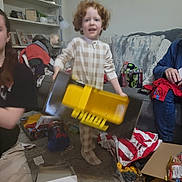 Kenji joined the competition — help win amazing prizes! child, toy, pajamas, living_room, couch, gift_wrap, adult, floor, box, shelf, plant, picture_frame, blanket, carpet, person, clothing, indoor, red_hair, smile, holiday