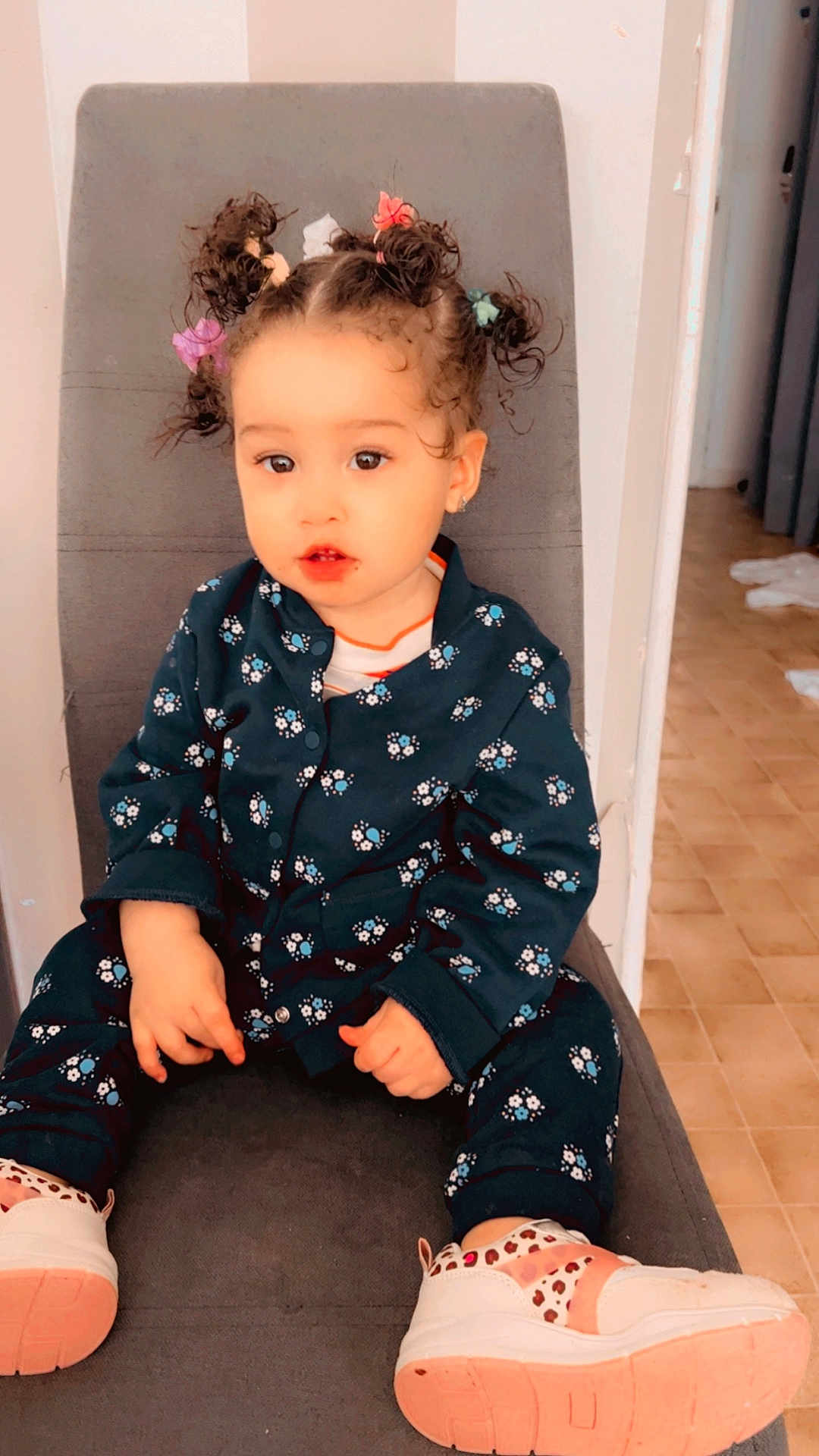 Catalina participe au concours pour gagner de l'argent avec cette photo : casual_wear, chair, child, curly_hair, cute, expression, floor_tiles, floral_clothing, footwear, hair_ties, home, indoor, light, person, portrait, seated, small_hands, sneakers, toddler, young_child