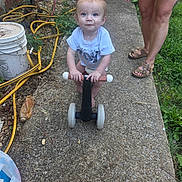 Jacob is registered to the contest to win money with this photo: toddler, child, baby, outdoor, garden, scooter, toy, sitting, standing, blue_shirt, smiling, face, portrait, adult, sandals, sidewalk, concrete, garden_hose, bucket, backyard
