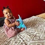 Khadija a rejoint le concours — aidez-le/la à gagner de superbes lots ! baby, child, plush_toy, bed, patterned_bedspread, red_wall, smiling, cute, indoors, pink_dress, person, soft_toy, happy, young, seated, portrait, skin, hair, baby_hair, cozy