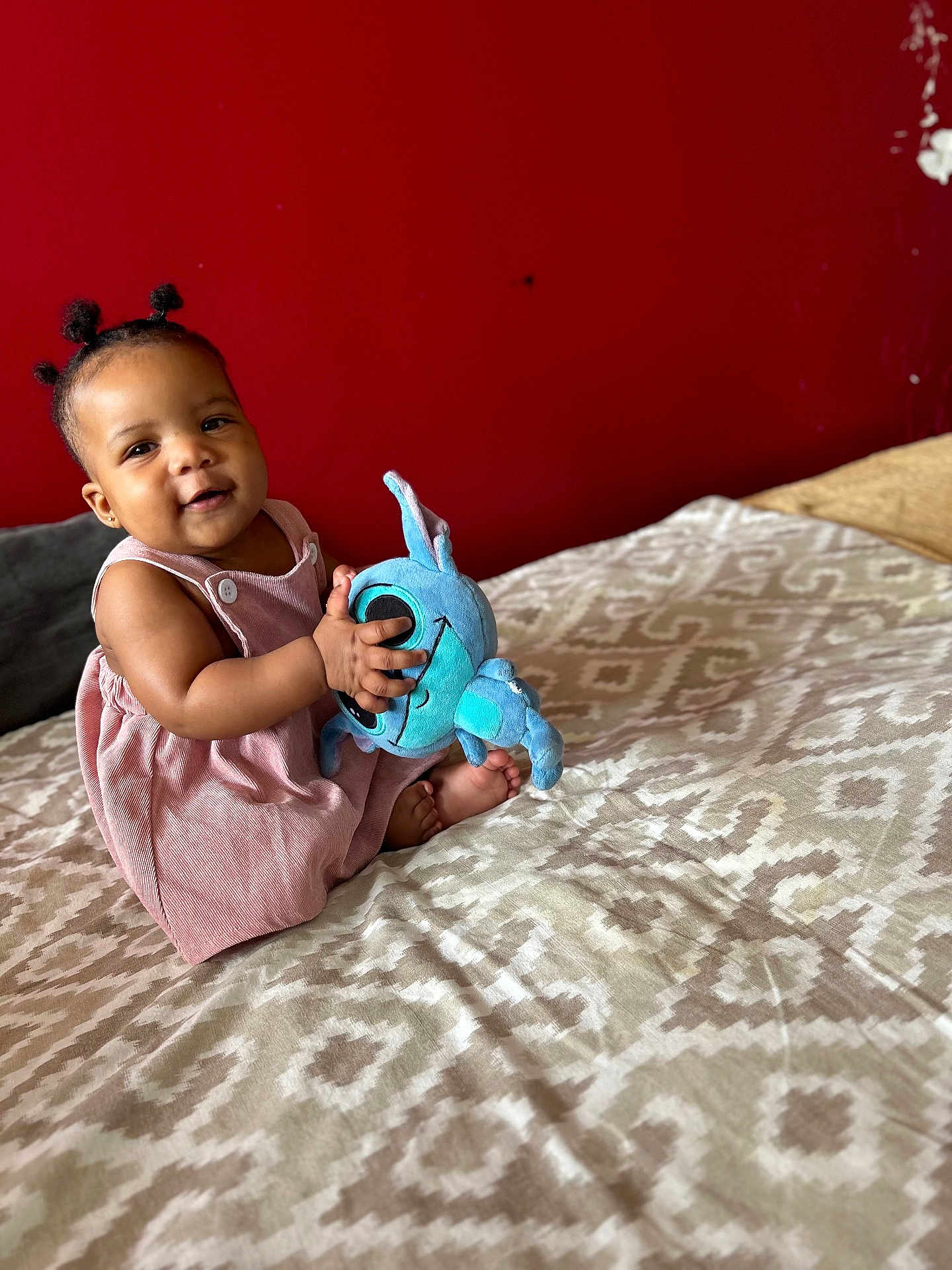 Khadija a rejoint le concours — aidez-le/la à gagner de superbes lots ! baby, child, plush_toy, bed, patterned_bedspread, red_wall, smiling, cute, indoors, pink_dress, person, soft_toy, happy, young, seated, portrait, skin, hair, baby_hair, cozy