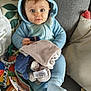 baby, infant, child, blue_onesie, hood, stuffed_animal, soft_toy, sofa, cushion, blanket, toy_book, teething_toy, beads, wide_eyes, sitting, portrait, indoor, cozy, plush, face