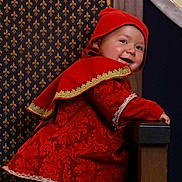 Tymaël a rejoint le concours — aidez-le/la à gagner de superbes lots ! baby, child, smiling, costume, red_clothing, hood, chair, patterned_wall, portrait, indoor, cute, happy, young_child, furniture, fabric, decoration, playful, person, headwear, footwear