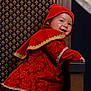 baby, child, smiling, costume, red_clothing, hood, chair, patterned_wall, portrait, indoor, cute, happy, young_child, furniture, fabric, decoration, playful, person, headwear, footwear
