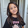 child, girl, smile, long_hair, black_tshirt, cartoon_characters, indoor, door, necklace, earrings, person, portrait, casual_clothing, cute, young, standing, happy, light, face, hair