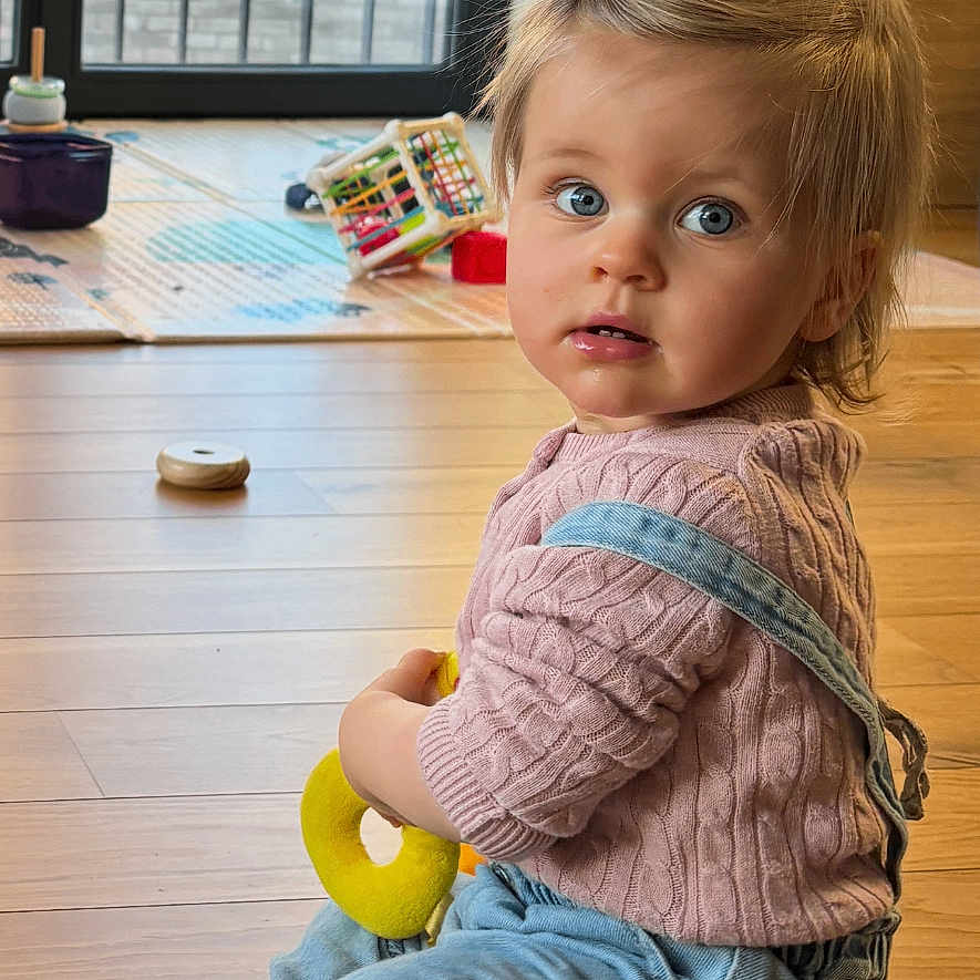 Elisabeth Mormede a rejoint le concours — aidez-le/la à gagner de superbes lots ! toddler, child, indoor, wooden_floor, toy, pink_sweater, blue_eyes, curly_hair, casual_clothing, sitting, playtime, daylight, home, window, flooring, denim_overalls, baby, cute, young_child, curious