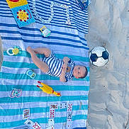 Naël Chamssedine participe au concours pour gagner de l'argent avec cette photo : baby, beach, sand, striped_towel, dominoes, playing_cards, soccer_ball, toy_chicken, toy_rattle, toy_mirror, baby_clothes, outdoor, child, blue_stripes, white_stripes, footwear_absent, sunny, relaxation, playtime, summer
