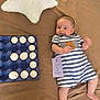 baby, infant, striped_clothing, egg_carton, eggs, number_three, card, pillow, star_shaped_pillow, brown_bedspread, smiling, lying_down, cute, celebration, indoors, child, face, hand, foot, happy