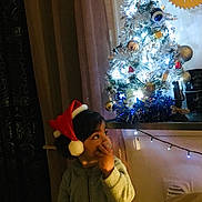 Miron a rejoint le concours — aidez-le/la à gagner de superbes lots ! child, toddler, santa_hat, christmas_tree, holiday, festive, decorations, lights, indoors, cozy, curtain, face, person, winter, seasonal, home, cute, young_child, soft_lighting, celebration