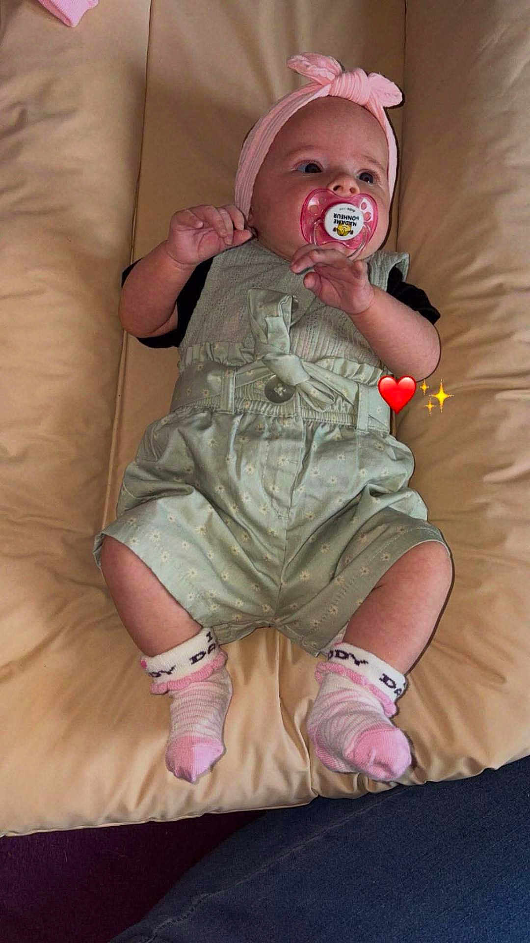 Célina-Marie participe au concours pour gagner de l'argent avec cette photo : baby, infant, pacifier, headband, pink, socks, cushion, clothing, cute, child, person, lying_down, indoors, soft_fabric, green_outfit, adorable, heart_emoji, sparkle_emoji, small_hands, relaxed