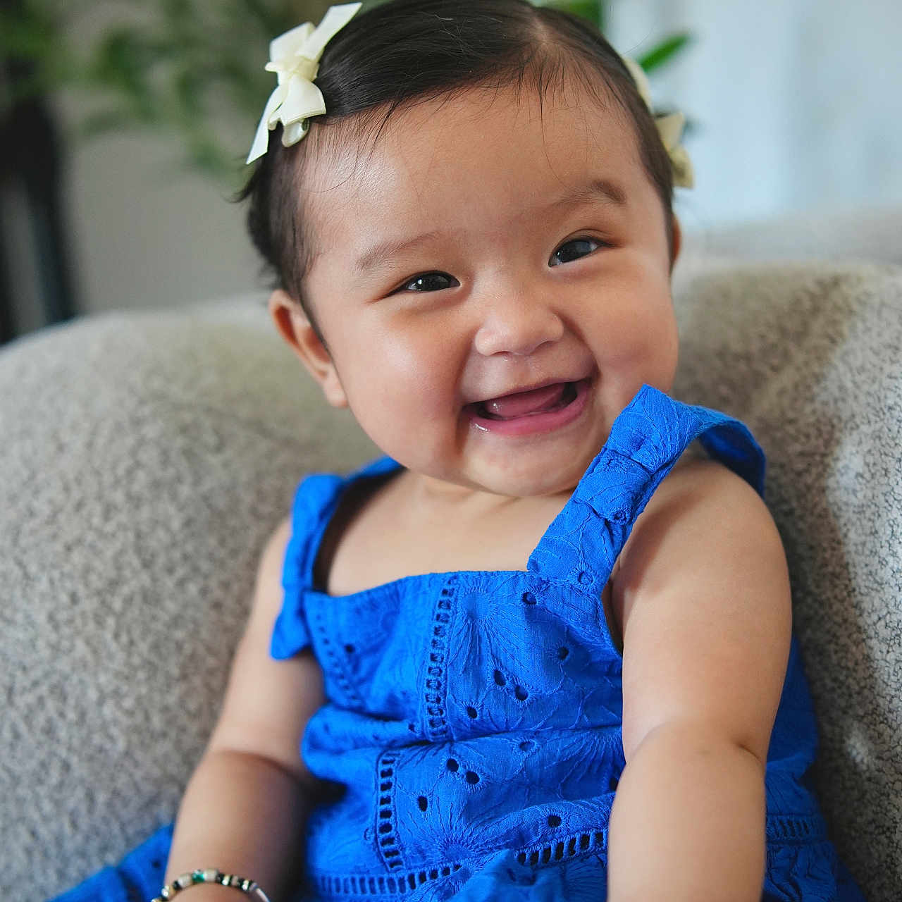 Zeya is registered to the contest to win money with this photo: baby, background_plant, blue_dress, bracelet, child, cozy, cute, expression, face, furniture, hair_bow, happy, indoor, person, portrait, sitting, skin, smiling, toddler, young_child