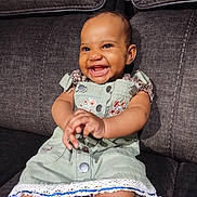 Seraphina is registered to the contest to win money with this photo: baby, smiling, child, couch, clothing, dress, happy, indoor, person, portrait, seated, cute, infant, hands, face, textile, fabric, button, embroidery, furniture