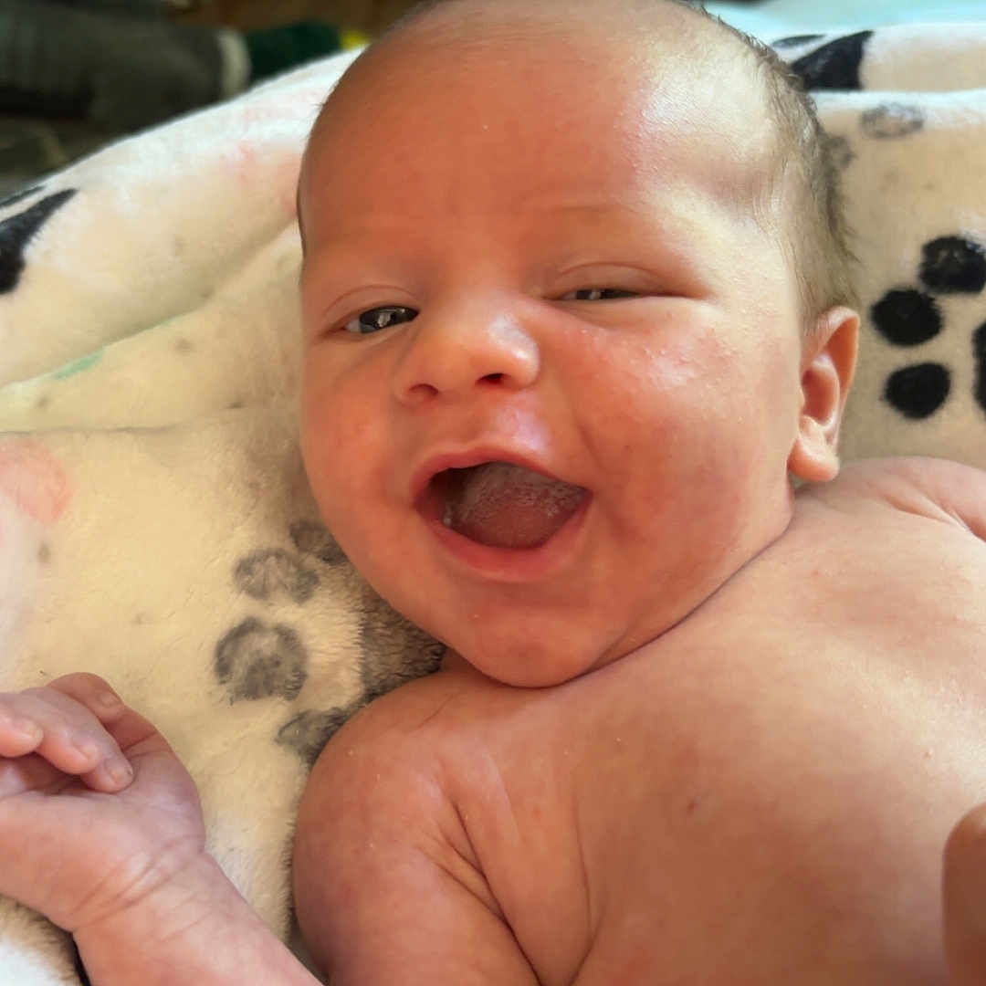 Trustyn is registered to the contest to win money with this photo: arm, baby, blanket, cheerful, child, closeup, cute, expression, face, happy, indoors, infant, lying_down, newborn, paw_print, person, portrait, skin, smiling, soft_texture