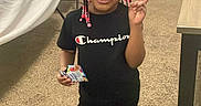 Amora joined the competition — help win amazing prizes! child, smiling, portrait, indoor, standing, peace_sign, braided_hair, hair_beads, black_shirt, champion_shirt, black_pants, sneakers, candy, snack, carpet, table, chair, curtains, birthday_decorations, family_room