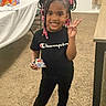 child, smiling, portrait, indoor, standing, peace_sign, braided_hair, hair_beads, black_shirt, champion_shirt, black_pants, sneakers, candy, snack, carpet, table, chair, curtains, birthday_decorations, family_room