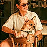 woman, person, french_bulldog, dog, pet, sunglasses, bow_tie, chair, wooden_chair, indoors, living_room, seated, portrait, fashion, smiling, brown_dog, animal, companion, home, warm_lighting