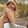 Danilo is registered to the contest to win money with this photo: baby, child, infant, bear_costume, hood, fluffy_clothing, outdoor, autumn, forest, trees, portrait, close_up, being_held, adult, hands, family, face, gazing, cute, golden_background