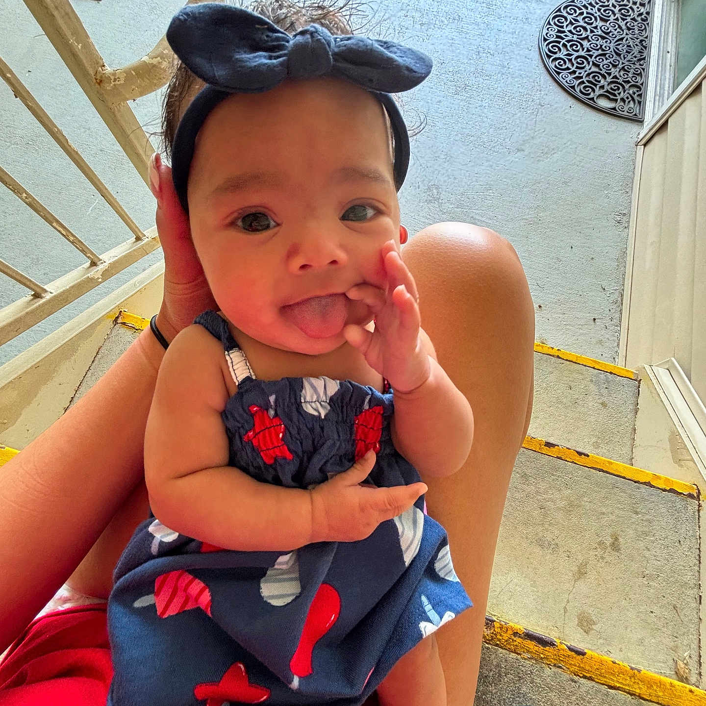 Khylan is registered to the contest to win money with this photo: adult, baby, blue_clothing, child, door, doormat, dress, expression, footwear, hand, headband, outdoor, person, playful, porch, red_clothing, sitting, skin, stairs, tongue_out