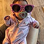 baby, infant, pacifier, sunglasses, pink_clothing, onesie, hand, lap, flooring, person, cute, indoor, child, feet, sitting, casual, relaxed, accessory, human, portrait