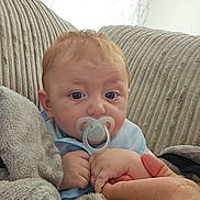Ayden is registered to the contest to win money with this photo: baby, pacifier, blanket, couch, striped_cushion, hand, person, indoor, family_photo, wall, patterned_wallpaper, blue_clothing, skin, face, infant, comfort, soft_texture, closeup, expression, holding_hand