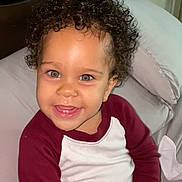 Hunter is registered to the contest to win money with this photo: toddler, child, curly_hair, smiling, bed, pillow, clothing, maroon, white, snack, happy, indoor, person, young_child, cute, portrait, sitting, face, eyes, cozy