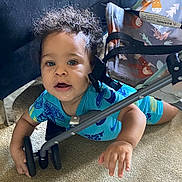 Hunter is registered to the contest to win money with this photo: toddler, child, baby, curly_hair, blue_clothing, crawling, stroller, wheel, carpet, indoor, hand, face, expression, curious, person, young_child, play, floor, bright_eyes, casual