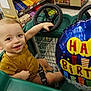 toddler, child, shopping_cart, balloon, birthday, smiling, store, books, candy, green, steering_wheel, clothing, floor, happy, person, indoor, seatbelt, baby, retail, celebration