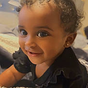 Laylah joined the competition — help win amazing prizes! toddler, child, smile, curly_hair, bows, black_clothing, blanket, bedroom, headboard, earrings, happy, indoors, portrait, cute, infant, baby, person, face, young_child, cozy