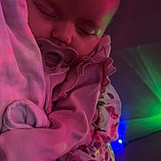 Vivian is registered to the contest to win money with this photo: baby, infant, pacifier, sleeping, blanket, swaddle, onesie, floral_pattern, ruffle, nightlight, colored_lights, purple_lighting, cozy, portrait, close_up, face, head, sleepwear, bedding, arm