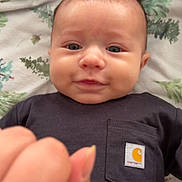 Daxton is registered to the contest to win money with this photo: baby, infant, face, smile, hand, black_shirt, logo, blanket, pattern, blue_eyes, skin, person, lying_down, cute, young_child, clothing, closeup, indoor, expression, playful