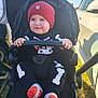 baby, child, stroller, red_hat, black_clothing, skeleton_outfit, smiling, outdoor, sunlight, grass, person, footwear, cute, infant, holding_bar, winter_clothing, jogger_stroller, casual, daytime, happy